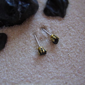 May include: Two black gemstone earrings with gold settings on silver posts. The earrings are on a white sandy surface.
