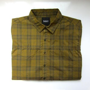 May include: A button-down shirt in a plaid pattern, featuring shades of olive green and dark gray. The shirt has a collar and a small black label with white text. The shirt is folded neatly.
