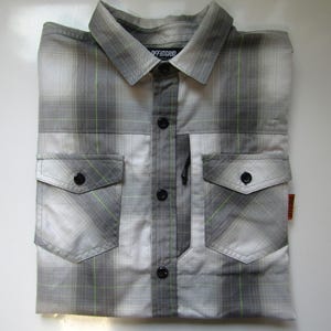 May include: A gray and white plaid button-up shirt with a black zippered pocket on the left side. The shirt has two flap pockets on the front and the text "OFF GRID" on the collar.