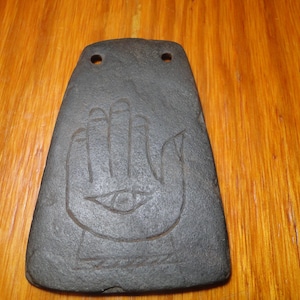 May include: A dark gray, trapezoidal stone plaque featuring a carved hand symbol with an eye centered in the palm. The object has two circular holes drilled near the top edge, providing a clear view of the textured, matte surface of the stone.
