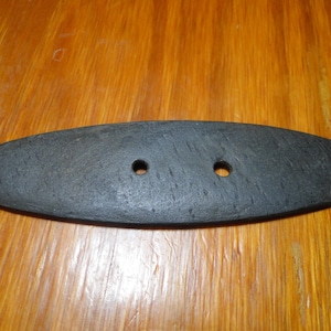 May include: A flat, elongated, oval-shaped black object with a textured surface and two circular holes positioned horizontally in the center. The piece has rounded ends and a matte finish, appearing as a hardware component or a decorative fastener.