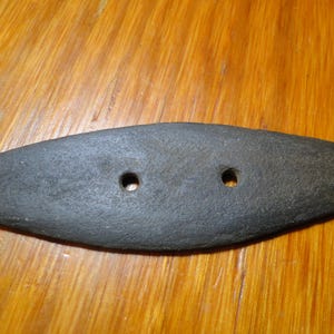 May include: A dark gray, elongated, oval-shaped object with a rough, textured surface. The item features two small, circular holes positioned horizontally in the center, creating a symmetrical appearance across its length.