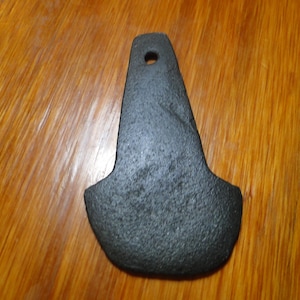 May include: A dark gray, flat, wedge-shaped stone object with a textured, porous surface. The object features a rounded, wide base that tapers toward a narrower top, which contains a single, circular hole centered near the upper edge.
