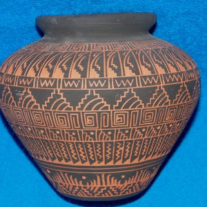 May include: A matte black ceramic pot featuring a wide, rounded body and a short, flared rim. The exterior is decorated with intricate, repeating geometric patterns in a terracotta orange color, including stepped lines, zigzags, and square spirals.