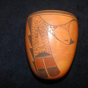 May include: A hand-painted, oval-shaped ceramic vessel with a light orange base. The vessel features a geometric design in dark brown and black, including triangles, rectangles, and dotted patterns. The rim is outlined in dark brown.