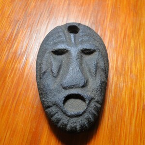 May include: A dark gray, oval-shaped mask with carved facial features. The mask has a prominent nose, open mouth, and eye sockets. A hole is at the top for hanging. The mask is likely made of stone or a similar material.