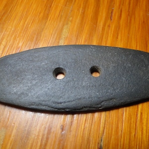 May include: An elongated, dark gray, flat stone button with a rough, natural texture. The button features two circular holes positioned in the center, designed for attachment to fabric or other materials through sewing or threading.