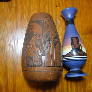 May include: A brown cylindrical vase with an etched Kokopelli figure and geometric patterns next to a smaller blue vase featuring a painted desert landscape with a mesa, horizontal black and white stripes, and a flared neck and base.