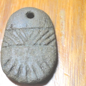 May include: A flat, oval-shaped gray stone pendant with a rough, textured surface. The stone features a drilled hole at the top for suspension and is carved with a series of radiating linear grooves that extend from the center toward the bottom edge.
