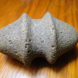 May include: A gray, textured stone artifact with a symmetrical, elongated shape featuring two prominent, rounded ridges encircling the center and tapered, rounded ends. The surface of the object is rough, porous, and speckled with darker mineral inclusions.
