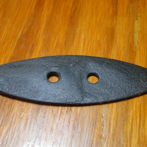 May include: A black, elongated, oval-shaped metal plate with a textured, matte finish. The object features two circular holes positioned horizontally along the center line, designed for mounting or attachment purposes with a flat, tapered profile.