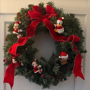 Pode incluir: Uma guirlanda de pinheiro artificial verde decorada com um grande laço de veludo vermelho, pequenas bagas vermelhas e pinhas. Quatro pequenas figuras de personagens da Disney com gorros de Papai Noel estão presas aos ramos.