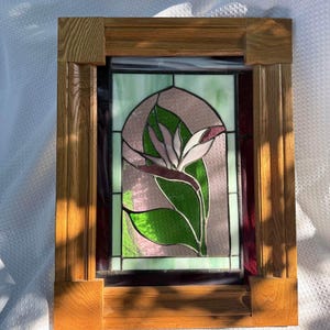 May include: A rectangular stained glass panel featuring a bird of paradise flower design in shades of green, white, and purple, encased within a thick, light-toned wooden frame with a visible grain pattern and a classic, sturdy construction.