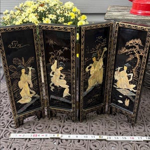 Puede incluir: Una pantalla plegable de cuatro paneles con acabado lacado negro y detalles dorados. Cada panel presenta figuras y paisajes asiáticos con incrustaciones de nácar. La pantalla mide aproximadamente 46 cm de ancho cuando está completamente extendida.