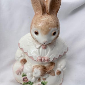 Peut inclure: Une figurine en céramique représentant un lapin beige et blanc portant un col blanc à bordure rose avec un petit nœud rose. Le lapin tient dans ses bras un plus petit lapin beige et blanc endormi, avec des fleurs roses et des feuilles vertes.