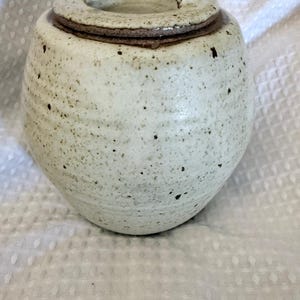 May include: A round, off-white ceramic jar with a speckled finish and a matching lid. The jar features a subtle horizontal ribbed texture along its body and a dark brown rim where the lid meets the base. The lid includes a small, arched handle.