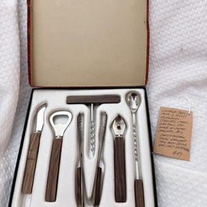 Op de afbeelding: Een zesdelige barset in een doos, bestaande uit roestvrijstalen gereedschappen met donkerhouten handvatten. De set bevat een mes, flesopener, tang, kurkentrekker, ijsprikker en een lange menglepel. Een handgeschreven label is bijgevoegd.