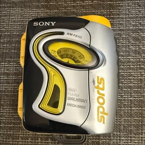 Puede incluir: Un reproductor de casetes Sony WM-FS111 Sports Walkman con carcasa de plástico negro, plateado y amarillo. El dispositivo incluye un dial de sintonización de radio analógico amarillo, etiquetas FM/AM, marca Mega Bass y la palabra sports en amarillo.
