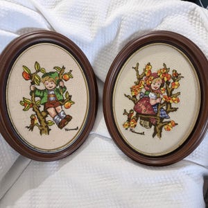 May include: Two oval wall hangings featuring embroidery on beige fabric within dark brown wooden frames. One depicts a child in a green jacket in an apple tree, the other a child in a red bonnet in a flowering tree. Both are signed Himmel.