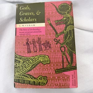 Peut inclure: Un livre relié intitulé Gods, Graves, & Scholars de C. W. Ceram. La couverture présente un fond rose et marron avec des illustrations noires de style égyptien, dont une figure à tête de chacal tenant un ankh et plusieurs petits personnages.