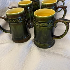 May include: A collection of six green ceramic beer mugs with brown handles and rims. Each mug features a decorative leaf band and a yellow interior. The mugs are displayed on a white surface.