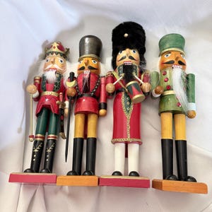 Peut inclure: Quatre figurines casse-noisettes en bois alignées. La première porte une couronne et tient une épée. La deuxième porte un haut chapeau et tient un bâton. La troisième porte un chapeau en fourrure noire et joue du tambour. La quatrième porte un chapeau vert.