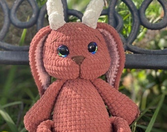 May include: A crocheted plush toy bunny with brown fur, white horns, and iridescent blue eyes. The bunny is sitting on a black metal bench.