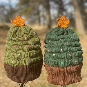 May include: Two knitted Christmas tree hats with brown brims and a star on top. One hat is green with colourful beads and the other is olive green with white beads.
