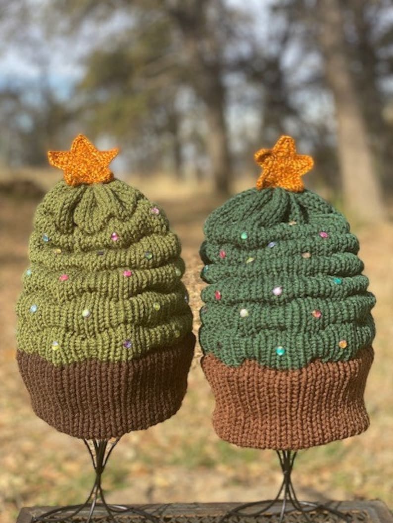 K&ouml;nnte beinhalten: Zwei gestrickte Weihnachtsbaum-M&uuml;tzen mit braunen Kanten und einem Stern oben. Eine M&uuml;tze ist gr&uuml;n mit bunten Perlen und die andere ist olivgr&uuml;n mit wei&szlig;en Perlen.