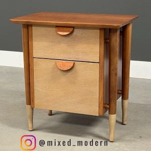 May include: A mid-century modern nightstand with two drawers. The nightstand is made of wood with a light brown finish on the drawers and a darker brown finish on the top and legs. The drawer pulls are half-circle shaped. The legs have a light-colored finish on the bottom.