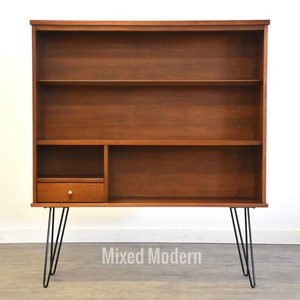 May include: A mid-century modern bookcase with three shelves and a drawer. The bookcase is made of wood and has black hairpin legs.