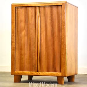 May include: A light brown wooden cabinet with vertical ribbed doors and a rectangular shape. The cabinet has a smooth, polished finish and sits on four short, angled legs. The doors have vertical grooves and a central handle.