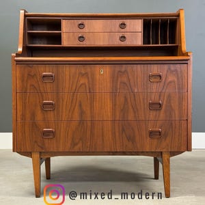May include: A mid-century modern wooden desk with a warm brown finish. It features three drawers, a small upper cabinet with two drawers, and open shelving. The desk has a keyhole and is supported by angled legs.