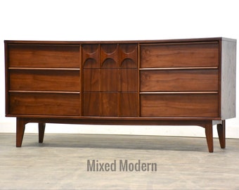 May include: A mid-century modern dresser with six drawers, featuring a unique geometric design on the centre drawer front. The dresser is made of dark wood and has tapered legs.