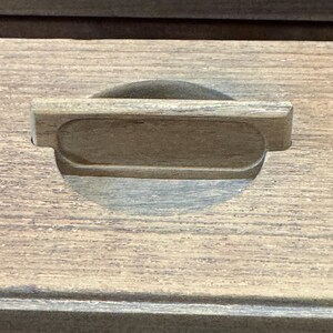 May include: Close-up of a wooden drawer front with a recessed handle. The handle is a rectangular shape with rounded edges, carved into the wood. The wood grain is visible, showing a natural, warm brown color. The drawer front has a simple, classic design.