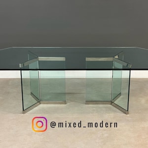 May include: A modern glass table with a rectangular top and geometric legs. The table is made of clear glass with a smoky tint and features stainless steel accents. The table measures approximately 72 inches long and 36 inches wide.