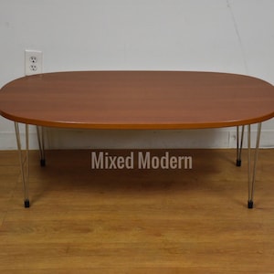 May include: A light brown oval coffee table with a wood top and chrome hairpin legs. The table is sitting on a wood floor.