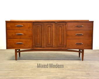 May include: Mid-century modern dresser with four drawers and two cabinet doors. The dresser is made of wood and has a light brown finish. The dresser has a unique design with a curved base and slender legs.