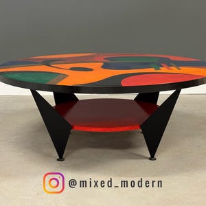 May include: A mid-century modern coffee table with a round top featuring an abstract design in orange, red, green, and black. The table has a black frame, a red lower shelf, and black angled legs. The Instagram handle @mixed_modern is visible.