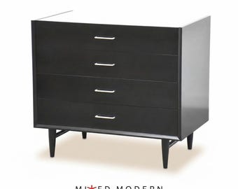 May include: A black four-drawer dresser with a white top surface and thin silver horizontal drawer pulls. The furniture piece stands on four tapered black legs and features the text MIXED MODERN with a red star icon below the base.