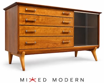 May include: A wooden sideboard featuring four horizontal drawers with rectangular handles and a glass-fronted cabinet section on the right side. The piece stands on four tapered legs and includes the text MIXED MODERN below it.
