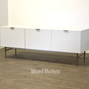 May include: White cabinet with three doors and gold metal legs. The cabinet has a textured diamond pattern on the doors. The cabinet is sitting on a wood floor.