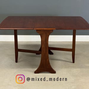 May include: A mid-century modern drop-leaf table with a rich, dark brown finish. The table features a unique pedestal base and slender legs. The tabletop is rectangular with rounded corners. The table is approximately 48 inches long when fully extended.