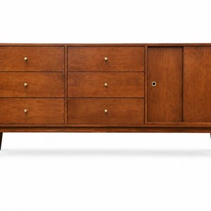 May include: A rectangular wooden dresser with a medium brown finish, featuring six drawers on the left and center with small round brass knobs, and a sliding cabinet door on the right with two circular brass pulls, supported by four tapered legs.