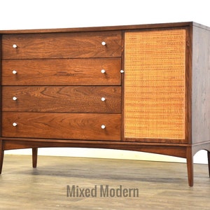 May include: A mid-century modern dresser with four drawers and a cabinet door with a woven rattan panel. The dresser is made of dark wood and has silver-toned drawer pulls.