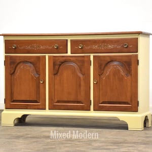 May include: A wooden cabinet with three doors and two drawers. The cabinet is painted in a light yellow color with a darker wood finish on the doors and drawers. The cabinet has a decorative design on the doors and drawers.