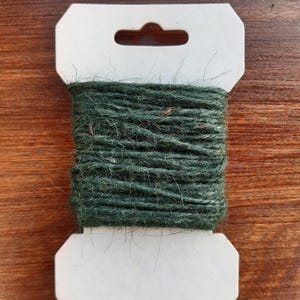 May include: A spool of dark green twine wrapped in a white cardboard holder with a hanging hole.