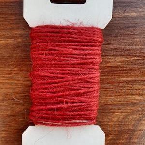 May include: A spool of red twine wrapped in white cardboard packaging with a hanging hole.