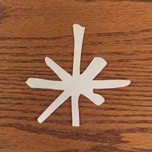 May include: A white paper snowflake cutout on a brown wooden surface.
