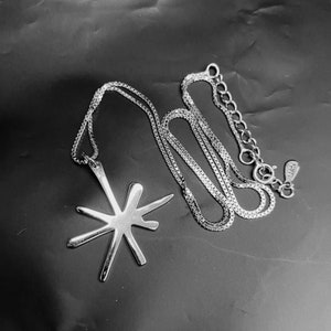 May include: A silver-toned necklace with a starburst pendant. The pendant has an abstract design with multiple radiating lines. The chain is a delicate box chain with an adjustable clasp. The necklace is displayed on a dark surface.